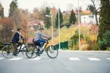 Two people wearing helmets ride electric bicycles across a pedestrian crossing on a quiet suburban street. They appear to be middle-aged and are dressed in autumn clothing. Trees with fall foliage and a few houses are visible in the background.